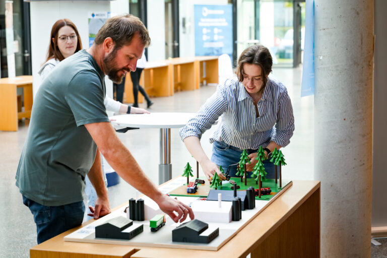 Ein Mann und eine Frau stehen vor einem Modell, dass auf einem Tisch steht. Es bildet die Lieferkette des Holzes ab: vom Forst bis zum Endverbraucher. Zu sehen sind Lagerhallen, Fahrzeuge und Bäume in der Größe von Playmobil Figuren. Im Hintergrund sieht man eine weitere Person.