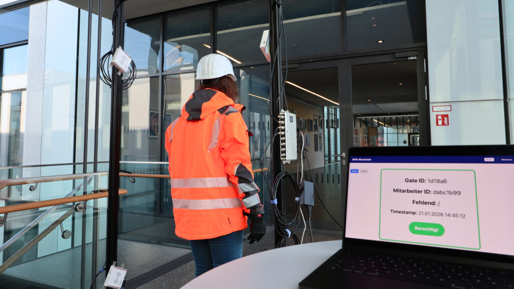 Zu sehen ist eine Frau von hinten. Sie trägt Arbeitskleidung, z.B. eine Orange Warn/-Arbeitsjacke und einen Helm, sowie Handschuhe. Sie geht durch ein Gate. Auf dem Bildschirm im Vordergrund wird eine Berechtigung angezeigt, dass alle Schutzkleidung vorhanden ist